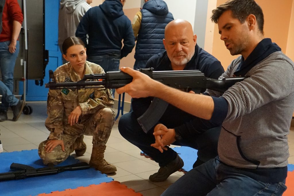 mężczyzna klęczy, trzyma karabin. obok kuca mężczyzna i dziewczyna w mundurze