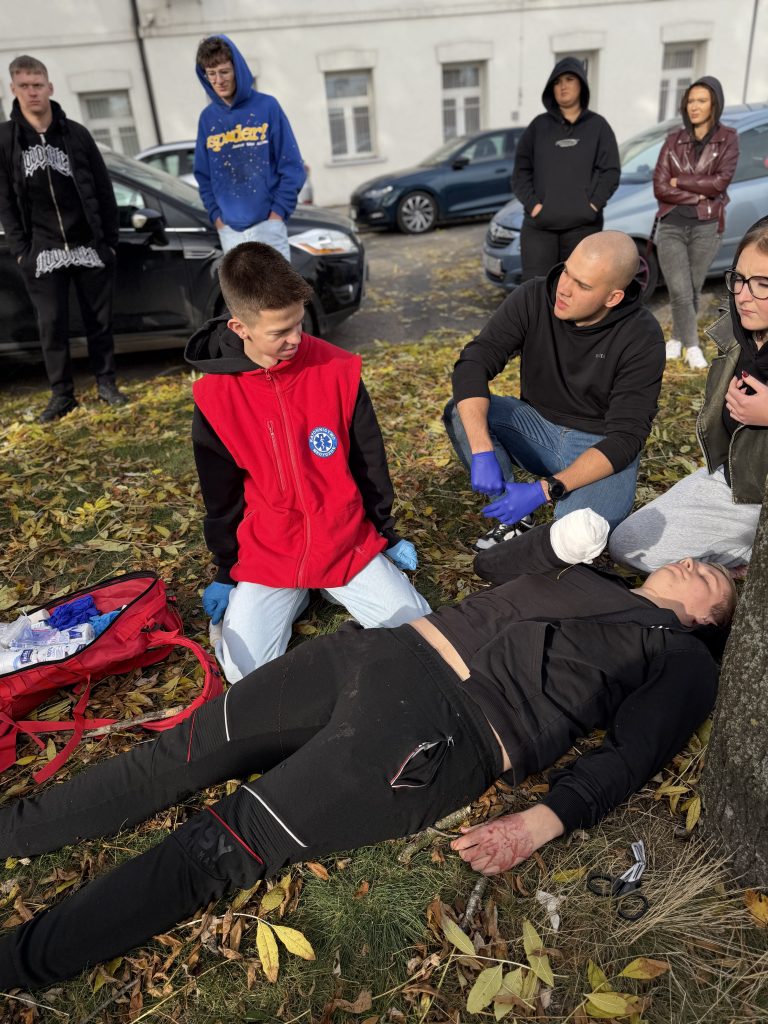 studenci sprawdzają stan poszkodowanego, który leży na trawie